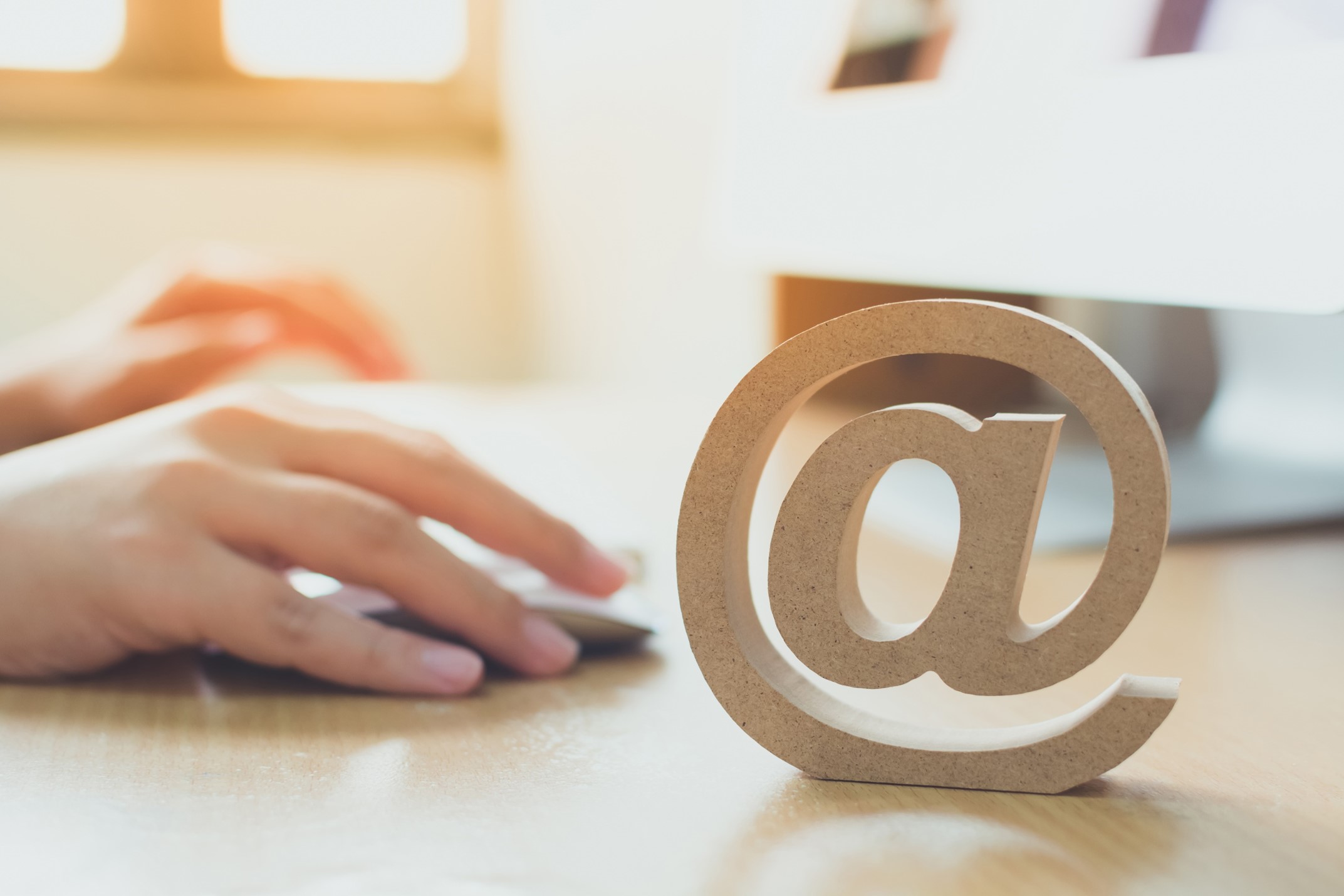 Close-up of a hand on a mouse beside a wooden at-symbol (@) representing email and digital workspace tools