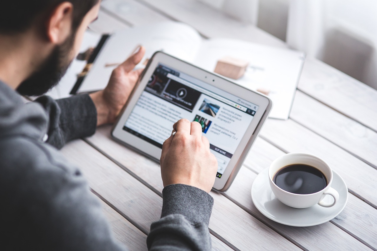 Person reading website content on a tablet with a stylus, sitting at a white table with a cup of coffee
